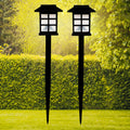 Two black solar garden lights with white paneled tops stand in a vibrant green lawn with hedges and trees.
